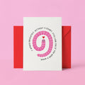 A white greeting card with a pink coiled worm illustration and black text reading 'I WOULD ABSOLUTELY WITHOUT A DOUBT STILL LOVE YOU IF YOU WERE A WORM' in reverse, shown with a red envelope against a pink background.