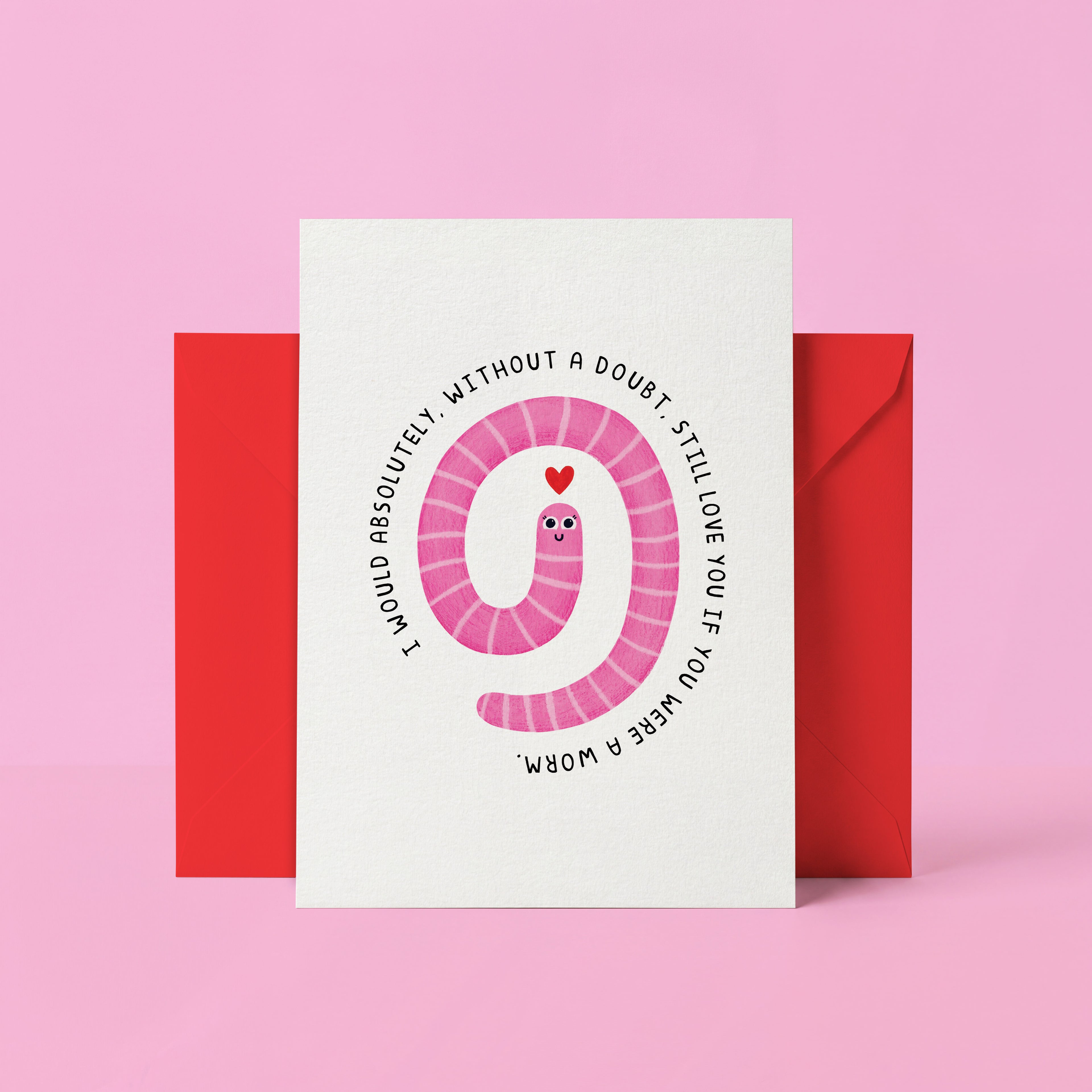A white greeting card with a pink coiled worm illustration and black text reading 'I WOULD ABSOLUTELY WITHOUT A DOUBT STILL LOVE YOU IF YOU WERE A WORM' in reverse, shown with a red envelope against a pink background.
